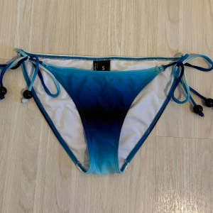 Blå bikiniunderdel med snörning från Twilfit - Snygg bikiniunderdel från Twilfit i blått med mörkare toning mot mitten. Knytband i sidorna med svarta pärlor ger en extra detalj. Perfekt för strandhäng och poolparty. Aldrig använt.