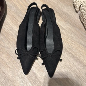 Svarta ballerinaskor från H&M med rosett - Svarta ballerinaskor från H&M i mockaimitation med spetsig tå och söt rosettdetalj framtill. Skorna har öppen häl och platt sula, vilket ger en stilren och trendig look. Perfekta för dig som gillar minimalistisk och chic stil. Använd 1 gång