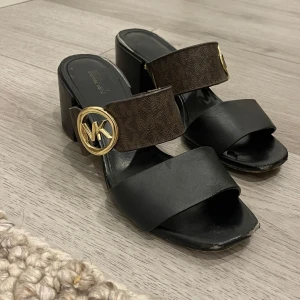 Svarta/bruna sandaler från Michael Kors - Sandaler med blockklack från Michael Kors med mörkbruna remmar och guldfärgad MK-logga på sidan. Tror att de är i storlek 37. Har lite skador vilket kan ses på bilden.