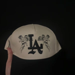 Sworn to us Vit LA keps med ängelbrodyr  - Snygg vit keps med svart LA-logga framtill och broderade änglar på sidorna. Justerbar snapback baktill för perfekt passform. Kepsen har coola svarta detaljer och är tillverkad i bomull för en skön känsla.