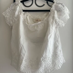 Vit blus med broderi från Hollister - Superfin vit blus från Hollister med broderade blommor och hålmönster. Blusen har korta puffärmar, resår vid halsringningen och romantiska spetsdetaljer längs kanterna. Perfekt för en somrig look. Skriv om du undrar något, kolla min profil innan du trycker på köp nu💘💘