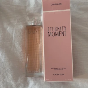 Calvin Klein Eternity Moment parfym - Eternity Moment från Calvin Klein är en elegant parfym i en rektangulär glasflaska med ljusrosa doftvätska. Flaskan har minimalistisk design och kommer med en matchande rosa kartong. Volymen är 100 ml eau de parfum spray. Aldrig använt