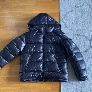 Svart Moncler Maya jacka - Säljer en svart, glansig pufferjacka från Moncler med huva och dragkedja framtill. Jackan har två snedställda fickor med dragkedja och klassisk Moncler-logga på ärmen. Perfekt för kalla dagar och riktigt snygg streetstil.