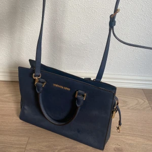 Mörkblå axelväska från Michael Kors - Snygg mörkblå axelväska från Michael Kors med guldiga metalldetaljer och dragkedja. Väskan har både handtag och justerbar axelrem, samt flera fack inuti för smart organisering. Tillverkad i tåligt skinn med stilren design och logga framtill.