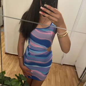 Blå och lila one shoulder kortklänning - Säljer en färgglad kortklänning med one shoulder och cut-out i midjan. Klänningen har vågiga ränder i blått, lila och rosa och sitter tight. Perfekt för dig som vill sticka ut med en trendig look!