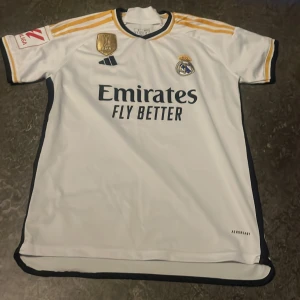 Real Madrid Bellingham tröja  - Real Madrid matchtröja med Bellingham 5 på ryggen. Vit med guld- och marinblå detaljer, Adidas-logga och FIFA World Champions 2022-märke. Klassisk rund hals med svarta och gula ränder, tillverkad i lätt polyester. Snygga gula ränder på axlarna och La Liga-märke på ärmen. Priset kan diskuteras 