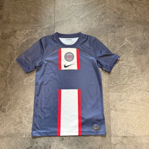 PSG matchtröja Nike - Paris Saint-Germain fotbollströja från Nike i marinblått med vita och röda vertikala detaljer på framsidan. PSG-logga och Nike Swoosh på bröstet. Tillverkad i lätt och ventilerande material, perfekt för träning eller match. Strl M i barnstrl