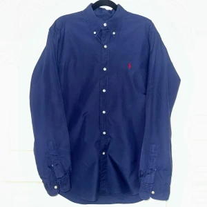 Mörkblå skjorta från Ralph Lauren M - Klassisk mörkblå skjorta från Ralph Lauren i bomull med button-down krage och vita knappar. Broderad röd logga på bröstet. Skjortan har lång ärm och en normal passform, perfekt till jeans eller chinos.