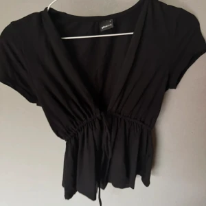 Svart omlottopp från Gina Tricot - Svart omlottopp från Gina Tricot med korta ärmar och knyt i midjan. Toppen har en djup v-ringning och en lätt volang nedtill som ger en snygg siluett. Perfekt att styla med jeans eller kjol för en trendig look.