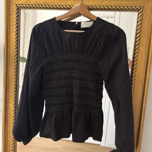 Svart blus med smock från Neo Noir - Supersnygg svart blus i bomull från Neo Noir med smockad midja och volang nertill. Blusen har långa ärmar med lätt puff och rund halsringning. Stängs med knapp i nacken. Perfekt för dig som gillar stilrena detaljer och en feminin siluett.