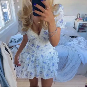 Blommig kortklänning med volanger - Supersöt kortklänning med blå blommor på vit botten, puffärmar och flera volanglager. Klänningen har v-ringning, spetsdetaljer och romantisk känsla – perfekt för sommarens alla tillfällen.