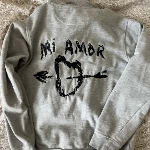 Grå hoodie Mi Amor från Mira Paris - Snygg grå hoodie från Mira Paris i mjuk ekologisk bomull. Baksidan har svart tryck med texten 'Mi Amor' och ett hjärta genomborrat av en pil. Klassisk känguruficka, snörning i huvan och relaxed fit. nypris är 1719 🩶