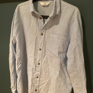 Ljusblå linneskjorta från H&M - Snygg ljusblå skjorta från H&M i luftig linneblandning. Klassisk krage, bröstficka och bruna knappar framtill. Perfekt för en chill och stilren look, passar lika bra till jeans som till shorts. Långärmad och med relaxed fit.