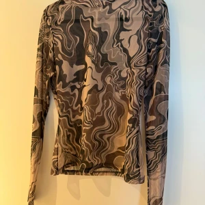 Mesh topp med camouflage-mönster - Säljer en tunn mesh topp med lång ärm och unikt camouflage-inspirerat mönster i beige, brunt och svart. Tight passform och genomskinligt material som ger en edgy vibe. Perfekt att styla lager på lager eller som statement-piece.