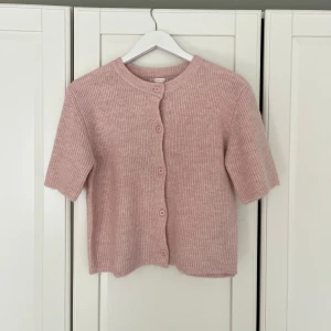 Ljusrosa stickad kofta med kort ärm - Superfin ljusrosa stickad kofta från Gina Tricot med korta ärmar och rund hals. Koftan har ribbad struktur och knäpps med matchande knappar framtill. Perfekt att slänga över en topp för en söt och chill look.