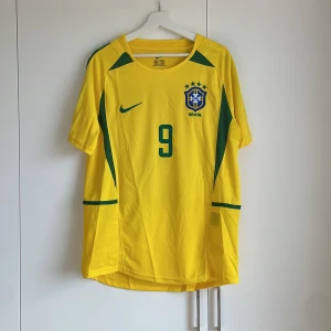 Brasilien Ronaldo 9 Nike fotbollströja M - Ikonisk Brasilien fotbollströja från Nike med nummer 9 och Ronaldo på ryggen. Tröjan är gul med gröna detaljer, kortärmad och tillverkad i lätt och ventilerande polyester. Perfekt för dig som älskar klassiska landslagströjor och vill sticka ut på planen.