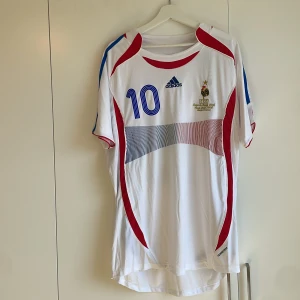 Frankrike Zidane 10 Adidas fotbollströja XL - Säljer en klassisk fransk landslagströja från Adidas med Zidane och nummer 10 på ryggen. Tröjan är vit med blå och röda detaljer, broderad tupp och stjärna på bröstet samt Adidas-logga. Kortärmad modell i lätt och ventilerande material, perfekt för fotbollsfans.