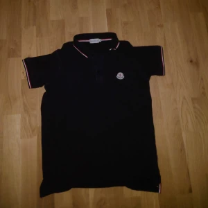 Svart Moncler pikétröja med logga - Svart pikétröja från Moncler i bomull med klassisk krage och knäppning framtill. Tröjan har korta ärmar och snygga rödvita detaljer på krage och ärmslut. Ikonisk Moncler-logga på bröstet. Perfekt för en clean och stilren look. Använd ungefär 7-10 gånger. Dma för fler bilder eller kvitto.