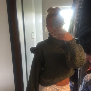 Grön stickad tröja med volang - Mysig mörkgrön stickad tröja med höga volangdetaljer på axlarna och ballongärmar. Tröjan har hög hals och är croppad i modellen, vilket ger en trendig och avslappnad vibe. Perfekt för kyliga dagar när du vill vara både bekväm och snygg.