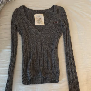 Grå stickad v-ringad tröja Hollister - Mysig grå stickad tröja från Hollister, vintage med v-ringning och lång ärm. Tröjan har ribbade muddar och ett Hollister-logo på bröstet. Snygg kabelstickad struktur och slim passform, xs men passar som s också!