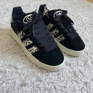 Adidas Campus sneakers svart/vit mönster - Snygga Adidas Campus sneakers i svart mocka med vita detaljer och coolt svartvitt mönster på de klassiska tre ränderna. Tjocka svarta skosnören, vit sula och vit häl med Adidas-logga. Perfekta för dig som vill sticka ut med en unik touch.