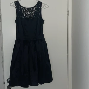 Svart spetsklänning från Anjou Glamour - Supersnygg svart kortklänning från Anjou Glamour med glansigt tyg och vacker spets upptill. Klänningen har ärmlös design, markerad midja och dragkedja bak. Perfekt för dig som vill sticka ut med en elegant look, eller använda vid fest/begravning eller valfria tillfällen! Säljer eftersom att den tyvärr blivit för liten.  Inga defekter, i nyskick och använd fåtal gånger. 