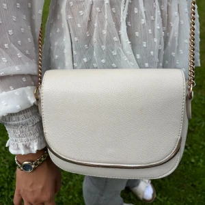 Vit axelväska med guldkedja - Jättefin vit axelväska i skinnimitation med guldfärgad kedja från HM. Väskan är använd men har ett bra skick. Skriv om ni har frågor💓