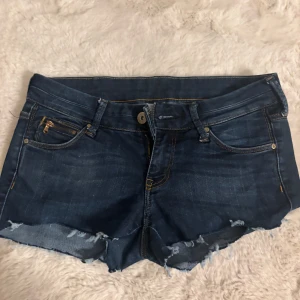 Mörkblå jeansshorts med fransar - Snygga mörkblå jeansshorts med råa, fransiga kanter och klassiska fickor fram och bak. Shortsen har guldfärgade detaljer på dragkedjan och knappen, samt slitningar för en cool, avslappnad look. Perfekta för varma dagar och festivaler.