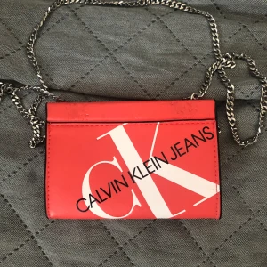 Orange plånbok / korthållare av  Calvin Klein Jeans - Snygg orange axelväska/ korthållare/ plånbok från Calvin Klein Jeans med stor vit CK-logga och svart text framtill. Väskan har en silverfärgad kedja och är tillverkad i slätt skinn. Perfekt storlek för det viktigaste och ger en cool statement-look.