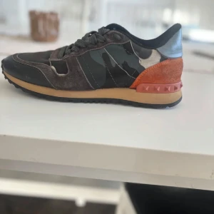 Valentino Rockrunner sneakers camo - Snygga Valentino Rockrunner sneakers med camouflagemönster i svart, grått och beige. Skorna har detaljer i mocka, skinn och orange mocka vid hälen med nitar. Sulan är beige och svart. Perfekt för dig som vill sticka ut med exklusiva sneakers. Storlek 39 passar 40. Inget og medföljer tyvärr. Skriv för minsta lilla fundering😁