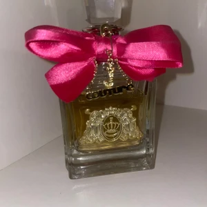 Juicy Couture parfym med rosa rosett - Juicy Couture parfym i en lyxig glasflaska med guldfärgad emblem och detaljer. Flaskan har en stor, glittrig kristallkork och är dekorerad med en iögonfallande rosa sidenrosett. Perfekt för dig som gillar glam och elegans. Tror de ca 70ml kvar av 100ml