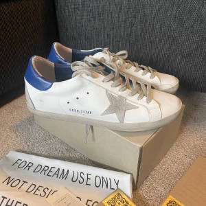 Golden Goose Super Star med Kvitto - Golden Goose Super Star sneakers i vitt skinn med grå mockadetaljer och signaturstjärna på sidan. Blå hälkappa i skinn, grå snörning och platt sula. Skorna följer med allt som nya, dvs kvitto, älthetsbevis, GG passport och dustbag. För mer bilder skicka dm.