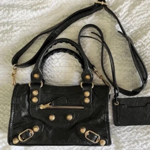 Svart handväska med gulddetaljer - Cool svart handväska i skinn med både handtag och axelrem. Väskan har snygga guldfärgade nitar, spännen och dragkedja framtill. Medföljer en matchande liten plånbok/korthållare. Perfekt för dig som gillar edgy detaljer och praktiska fack.