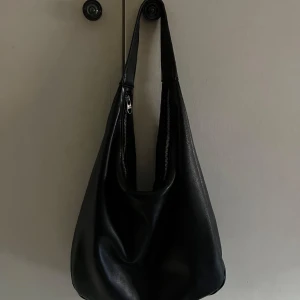 Svart axelväska i skinnlook - Stilren svart axelväska med mjuk form och bred axelrem. Väskan har en dragkedja och materialet är i skinn. Inuti finns ett litet fack som är perfekt för att förvara småsaker. En perfekt väska till vardagen som du får plats med allt du behöver i💕