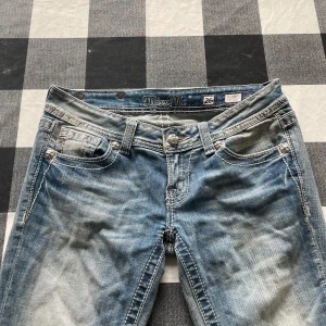 Miss Me ljusblå bootcut jeans med brodyr - Snygga ljusblå jeans från Miss Me med bootcut passform och slitna detaljer. Baksidan har broderade peace-märken på fickorna och coola nitar. Jeansen har klassisk femficksdesign och kontrastsömmar. Perfekta för dig som gillar statement-plagg! OBS, defekter vid jeansen men som ej är så märkbara när man har på sig dom. Annars i ett bra skick. Jag är 1,64