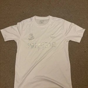 Vit Balmain x Adidas t-shirt x Real Madrid  - Snygg vit t-shirt från Balmain och Adidas collab med diskret glansigt tryck framtill. Klassisk rund hals och korta ärmar. Tillverkad i mjukt och lätt polyestermaterial som andas. Perfekt för dig som gillar stilrena och exklusiva plagg.