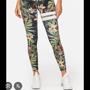 Blommiga leggings med tropiskt mönster - Snygga leggings med tropiskt blommönster i grönt, gult och rosa. De har två vita ränder på ena låret som cool detalj. Perfekta för dig som vill sticka ut på gymmet eller i vardagen. Stretchigt material som sitter tight och bekvämt.