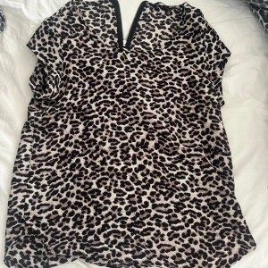 Leopardmönstrad blus med dragkedja - Snygg blus med leopardmönster i svart, beige och brunt. Toppen har korta ärmar och en cool dragkedja både fram och bak vid halsringningen. Perfekt för dig som vill sticka ut med ett djurmönster och gillar trendiga detaljer.