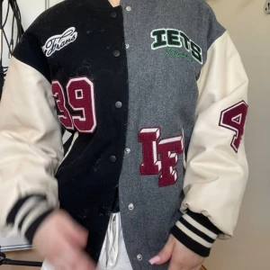 Collegejacka från Iets Frans, svart/grå - Snygg bomberjacka från Iets Frans med svart och grå paneler, krämvita ärmar och broderade patches i vinrött och grönt. Jackan har tryckknappar, randiga muddar och coola detaljer både fram och bak. Perfekt statement-plagg för dig som gillar streetstyle.