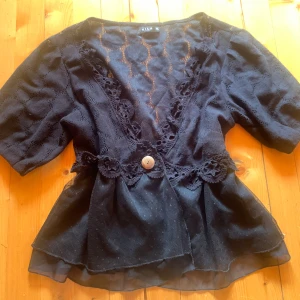 Svart spetsblus från Vila med volang - Svart blus från Vila med v-ringning, spetsdetaljer och blommiga broderier längs midjan. Är i storlek xl men passar mig som har s. Korta ärmar, volang nertill och en dekorativ knapp framtill. Tunn och luftig med transparenta partier och snygg struktur i tyget.