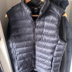 Svart dunväst från Uniqlo - Säljer en svart, quiltad dunväst från Uniqlo Ultra Light Down. Västen har hög krage, dragkedja framtill och är superlätt. Perfekt att slänga över en hoodie eller tröja när det är kyligt ute.