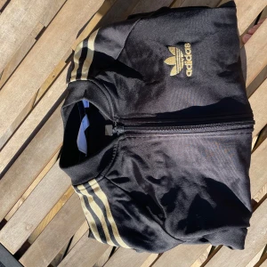 Svart Adidas bomberjacka med guld - Svart Adidas bomberjacka typ ziphoddie med klassiska guldiga ränder längs ärmarna och broderad logga på bröstet. Jackan har dragkedja framtill, ribbade muddar och två fickor. Perfekt för en sportig och stilren look.