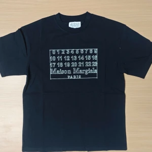 Maison Margiela svart t-shirt - Svart t-shirt från Maison Margiela med ikoniskt tryck framtill med siffror och logga. Klassisk passform, rund hals och korta ärmar. Tillverkad i mjuk bomull som känns skön mot huden. Perfekt för dig som gillar stilrena och coola plagg.