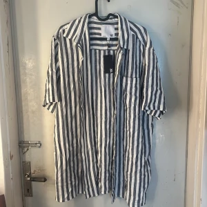 Randig skjorta  - Snygg skjorta från lager 157 i storlek XL, tillverkad i luftig linneblandning. Skjortan har vertikala svarta och vita ränder, kort ärm, klassisk krage och knäppning framtill. Perfekt för dig som gillar en avslappnad och trendig stil. Köpt för 150 kr