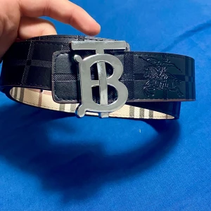 Rutmönstrat bälte från Burberry med silverfärgat spänne - Säljer ett snyggt bälte från Burberry med klassiskt rutmönster i svart och beige. Bältet har ett stort silverfärgat TB-spänne och är tillverkat i skinn. Perfekt accessoar för att lyfta din outfit.