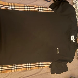 Svart T-shirt med rutiga sidopaneler från Burberry - Säljer en svart T-shirt från Burberry med klassiska beige, röda och svarta rutiga paneler längs sidorna. T-shirten har rund halsringning och korta ärmar. Perfekt för dig som gillar stilren design med en touch av ikoniskt mönster.
