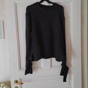 Mörkgrå stickad tröja med knyt vid ärmslut - Säljer en mörkgrå stickad tröja med långa ärmar och snygga knytband vid ärmsluten. Tröjan har en rund halsringning och en rak passform. Perfekt för kyligare dagar och enkel att matcha med olika outfits.