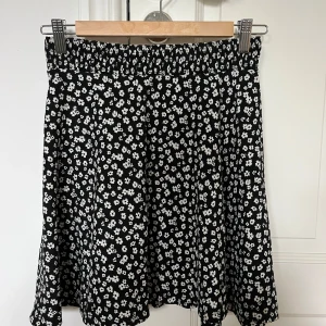 Svart blommig kortkjol - Supersöt svart kortkjol med vita små blommor över hela tyget. Kjolen har resår i midjan för extra komfort och en lätt utsvängd passform. Perfekt att styla med en enkel topp eller hoodie. A-Linje kjol.