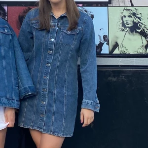 Jeansklänning med knappar - Säljer en klassisk jeansklänning i blå denim med långa ärmar och knappar framtill. Klänningen har en rak passform och sömdetaljer längs framsidan samt bröstfickor. Perfekt för dig som gillar en tidlös och avslappnad stil.