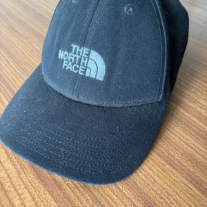 Svart keps från The North Face - Säljer en svart keps från The North Face med broderad logga framtill. Klassisk böjd skärm och justerbar passform baktill. Perfekt för en sportig och avslappnad stil.